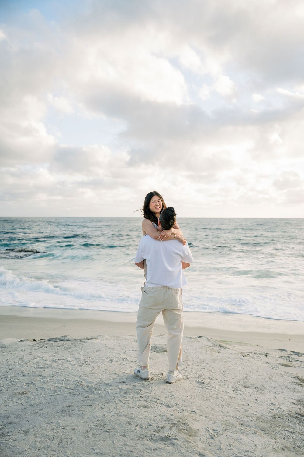 A romantic couple embrace on a sandy beach with crashing waves and cloudy skies in the background.