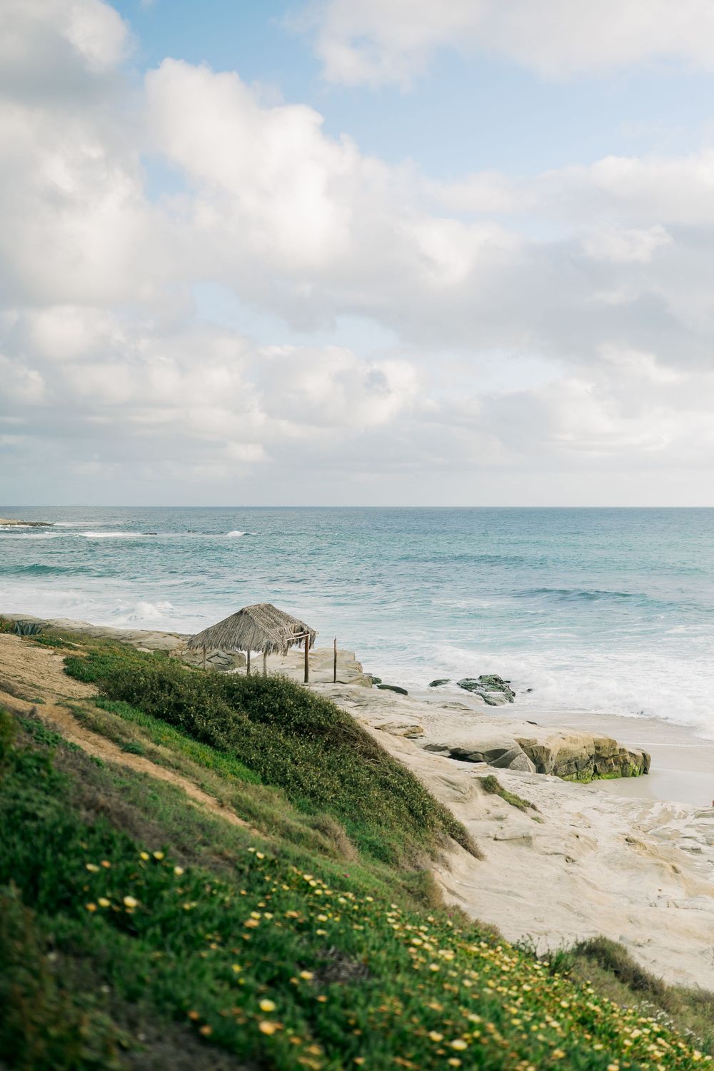 A scenic coastal landscape with a thatched beach hut perched on a grassy cliff overlooking turquoise waters and sandy beach.