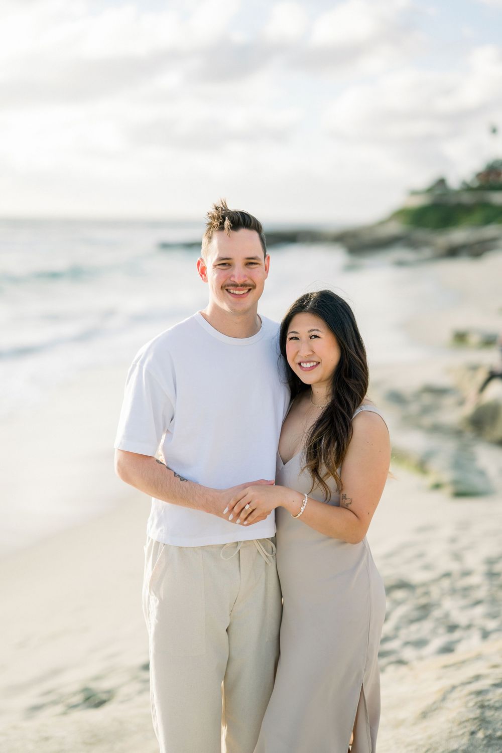 A couple in coordinated white and beige outfits share a romantic moment on a scenic beach at sunset.