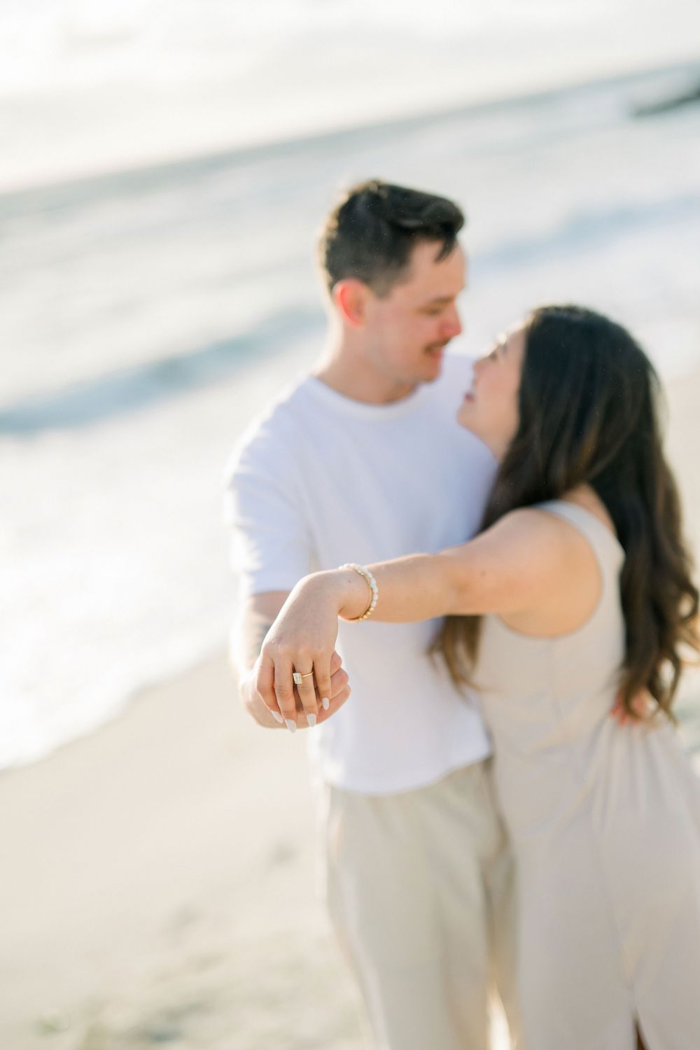 A romantic couple shares an intimate moment on a sunlit beach in coordinated white and neutral outfits.