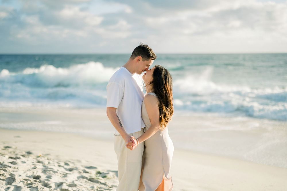 A couple shares an intimate moment while standing together on a sunny beach with ocean waves behind them.