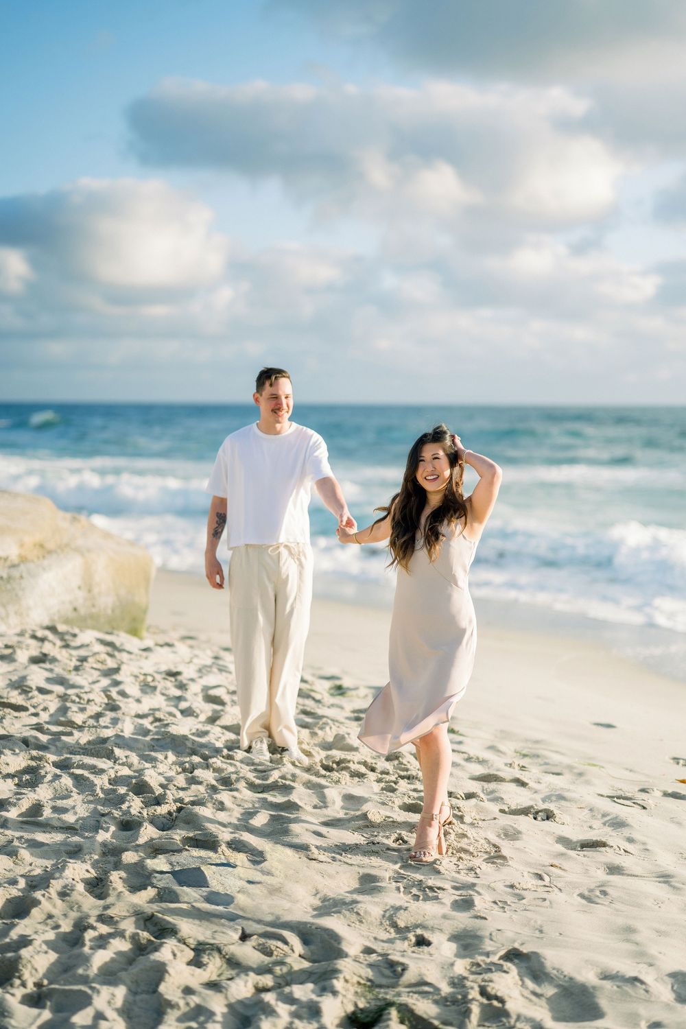A couple in white outfits walking and laughing together on a sunny beach with waves and blue sky in the background.