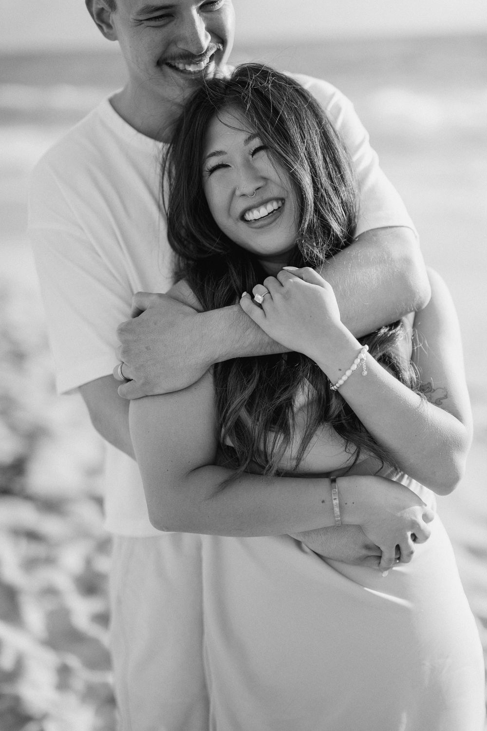 A couple embraces and laughs together on a sunny beach with ocean waves in the background.