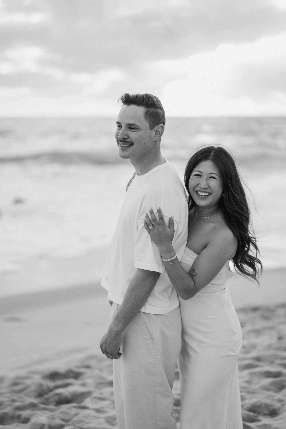 A happy couple in white outfits shares a candid moment together on a beach during a black and white photoshoot.