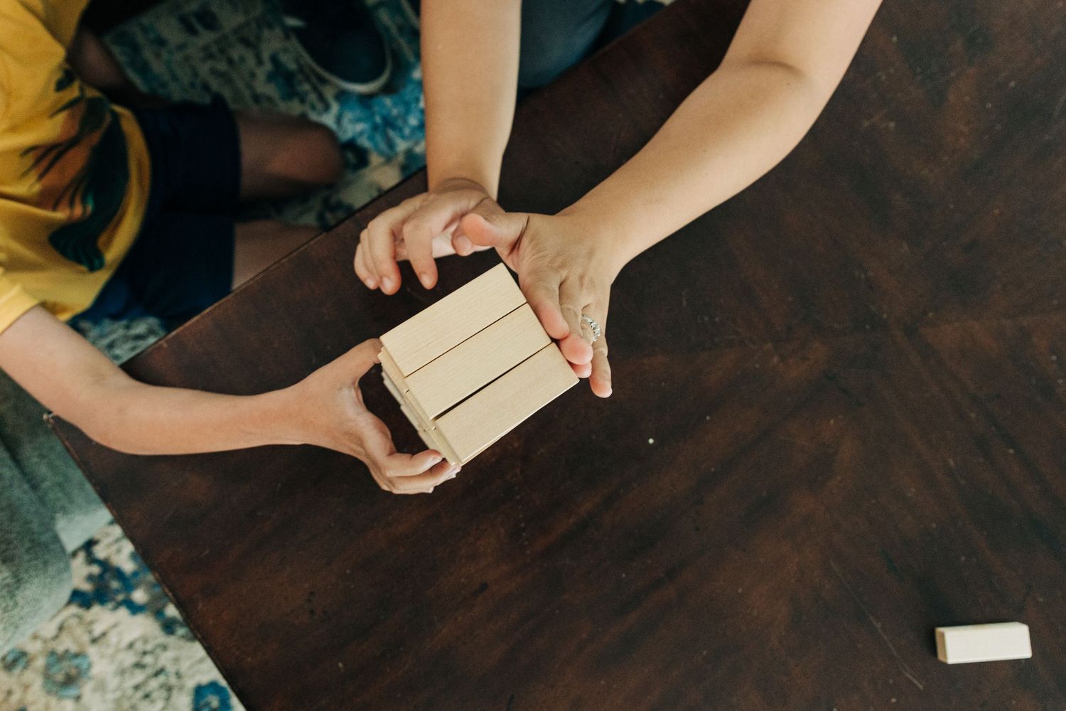 Hands exchanging a small white gift box on a dark wooden table surface with scattered blue decorative elements.