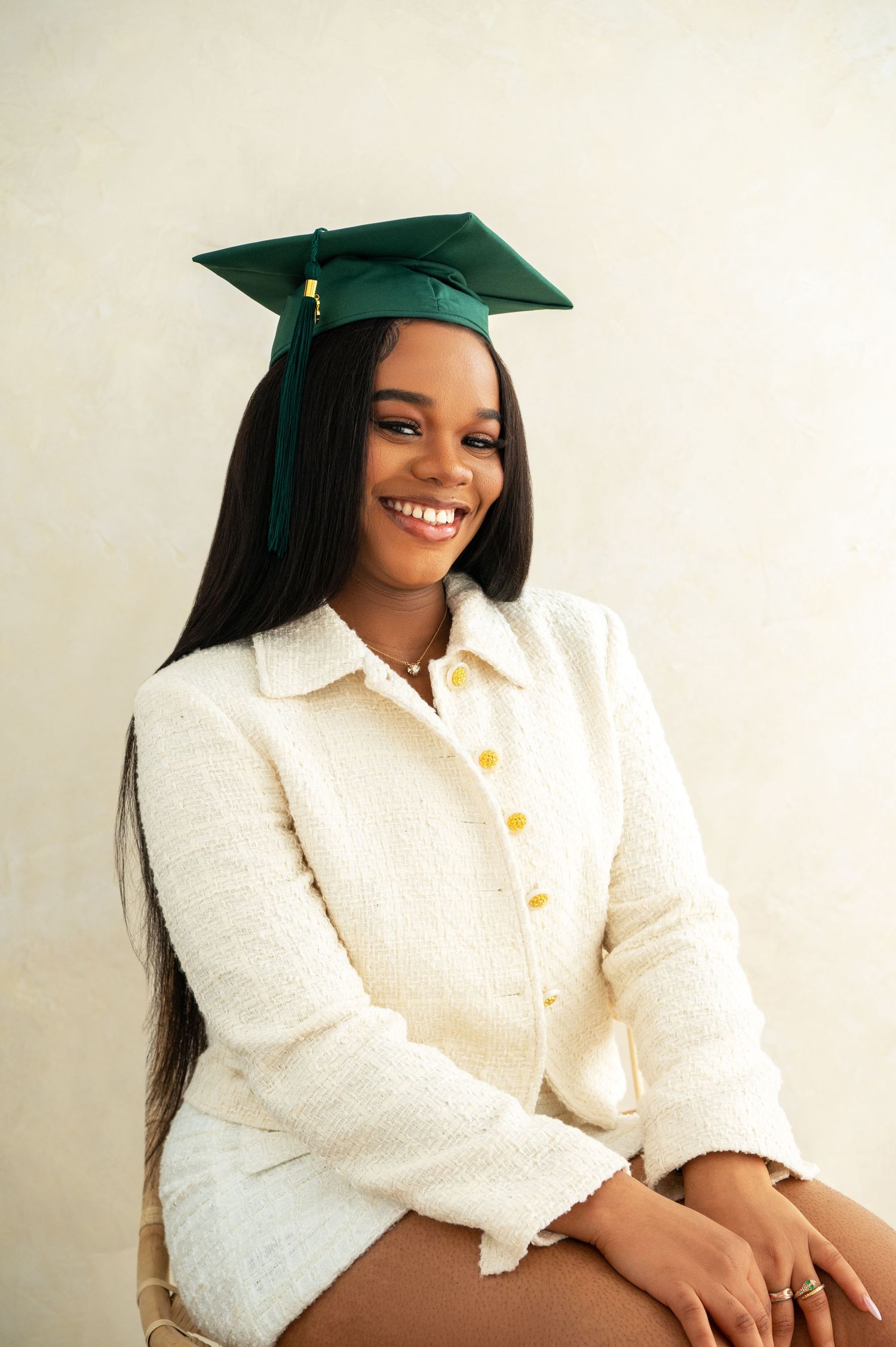Graduate in white sweater and green cap smiles during portrait session against beige wall.