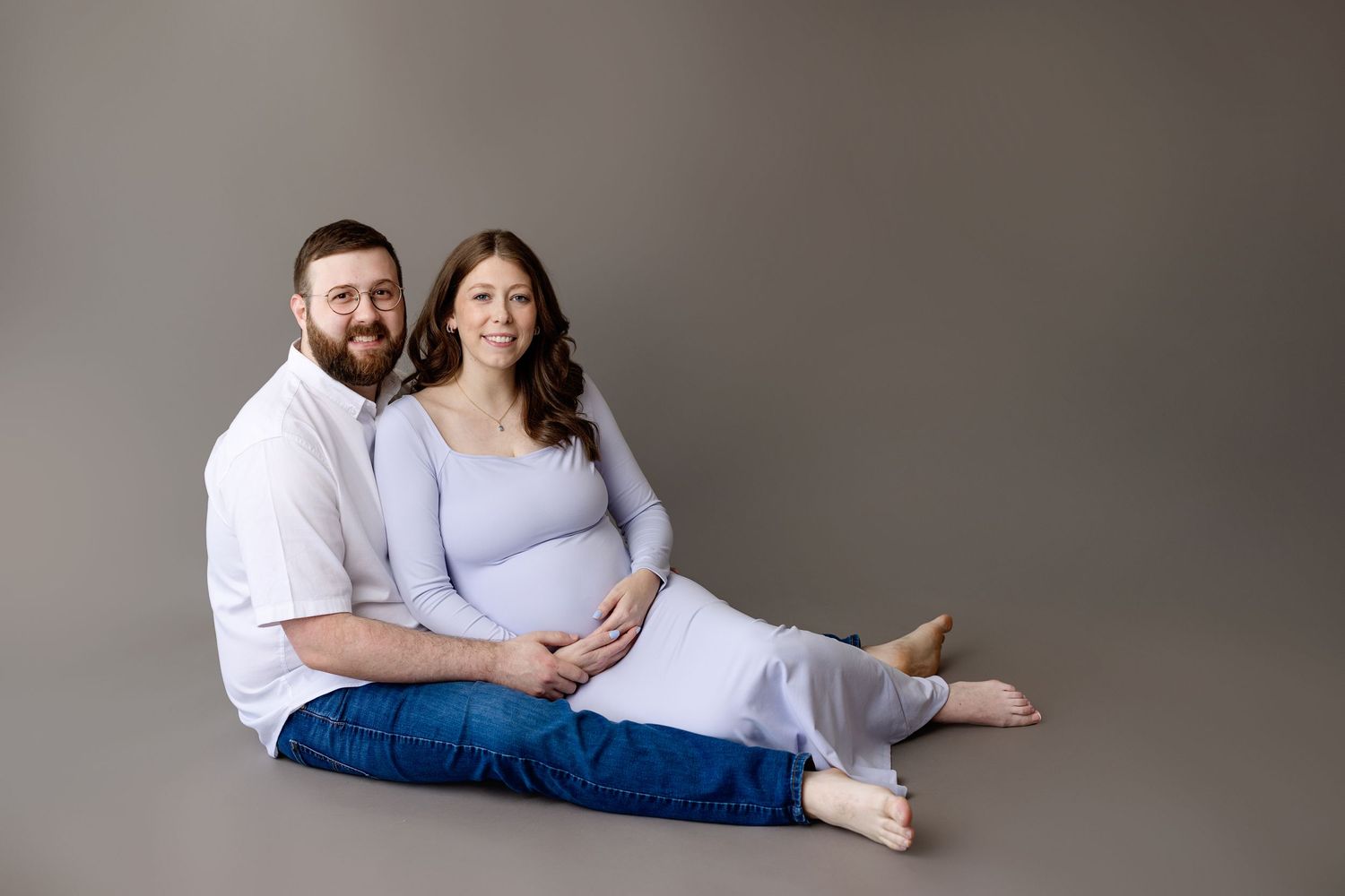 A couple sits together against a neutral grey backdrop wearing coordinated white and blue outfits for a studio portrait.