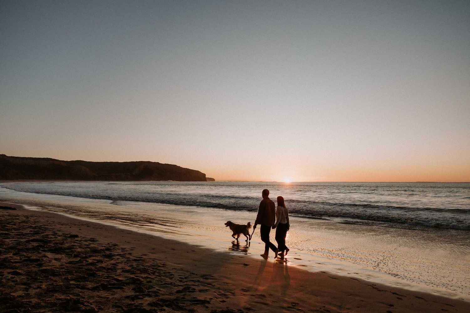 Silhouettes of people walking dog on sandy beach during golden sunset with cliffs and ocean waves in background.