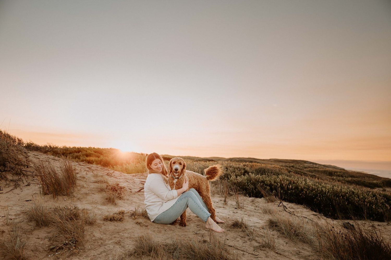 Person sits on sandy beach dunes with dog watching golden sunset over coastal hills on warm evening.