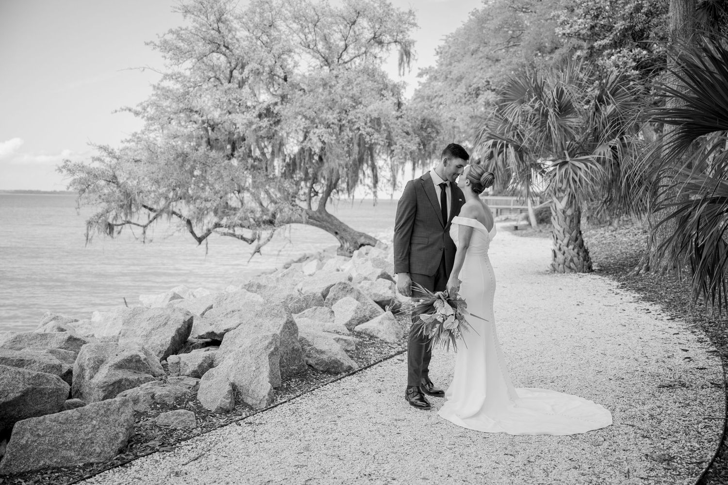 A couple shares a romantic moment on a coastal path surrounded by rocks, trees and palm fronds in black and white.