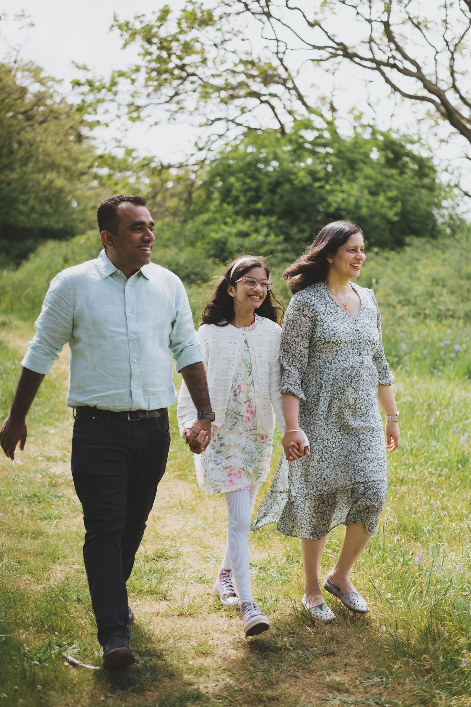 A family walks together holding hands on a dirt path through green grass and trees on a sunny day.