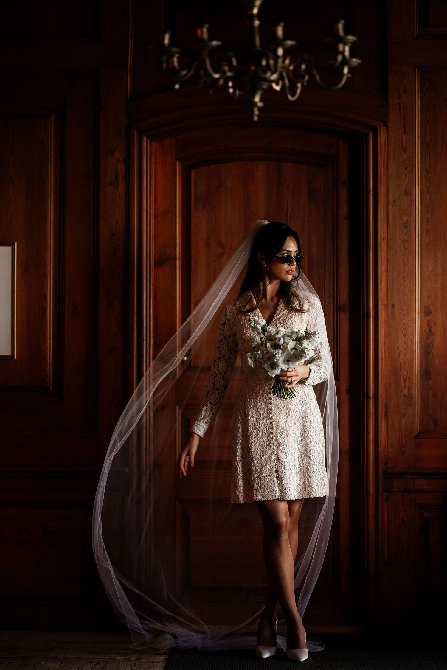 A bride in a short lace wedding dress and veil poses elegantly against dark wood paneling in moody lighting.