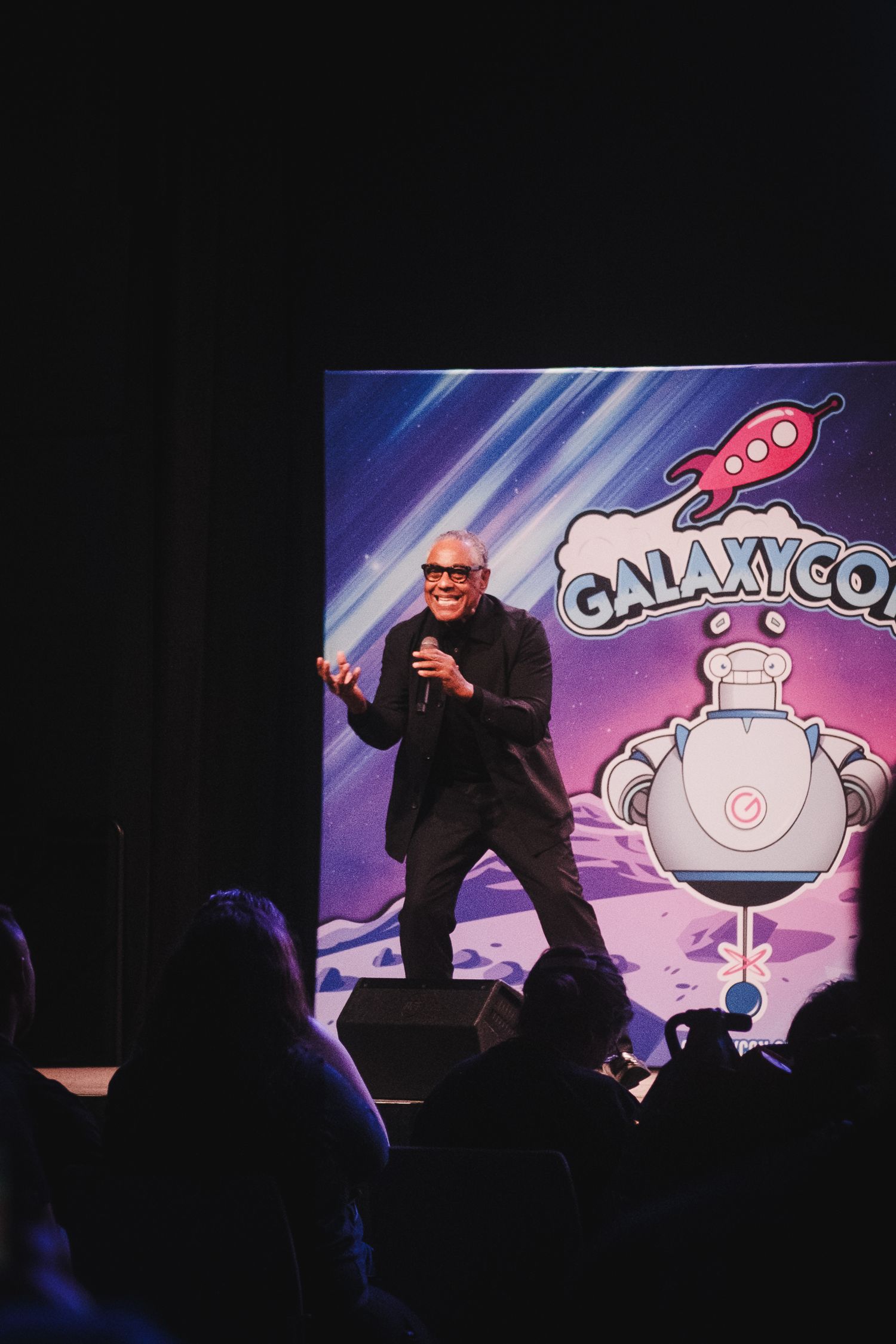 A performer stands on stage gesturing with a microphone in front of a colorful Galaxy Con backdrop with a cartoon robot design.