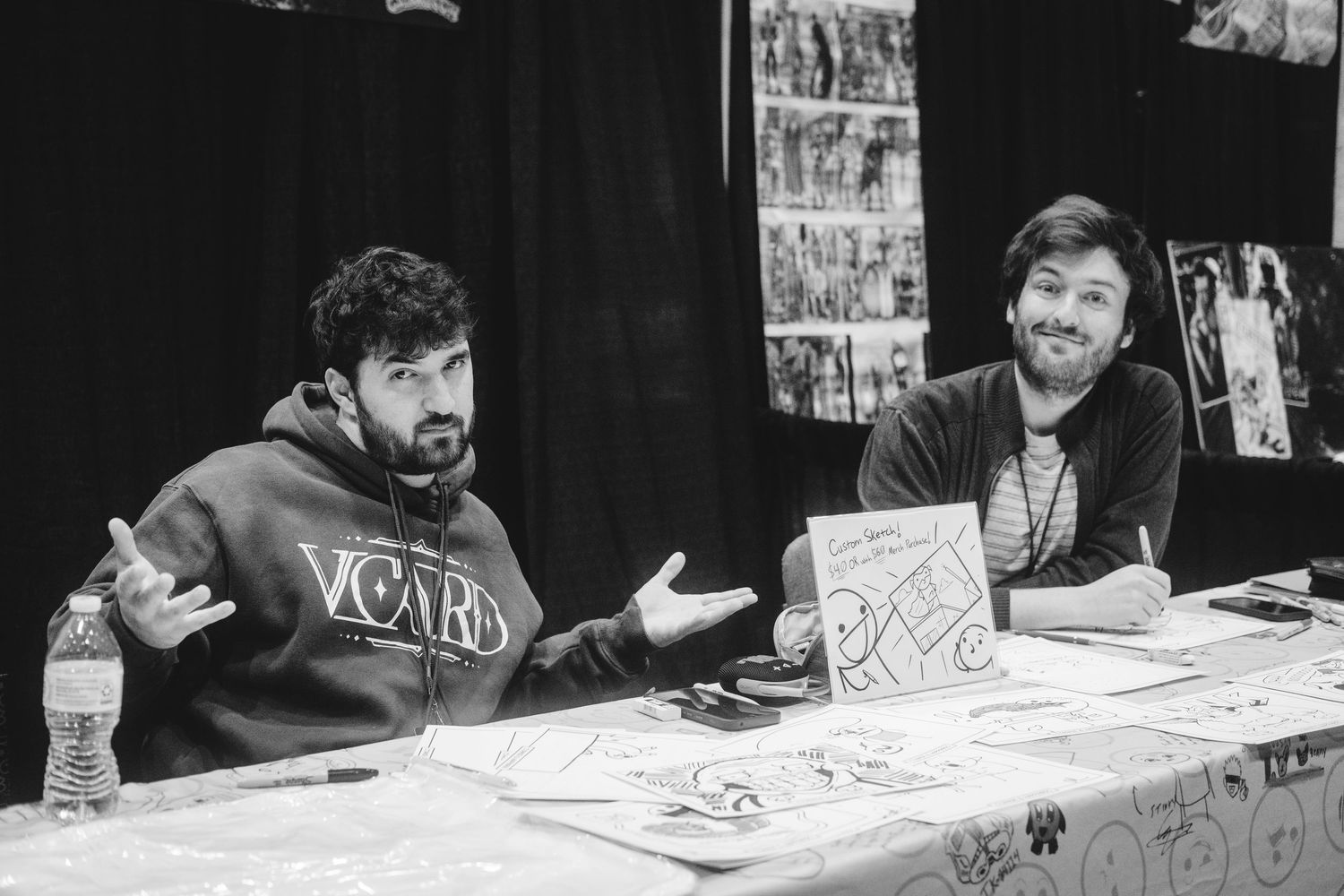 Two men sit at a signing table at what appears to be a convention, with papers and photos visible on the table.