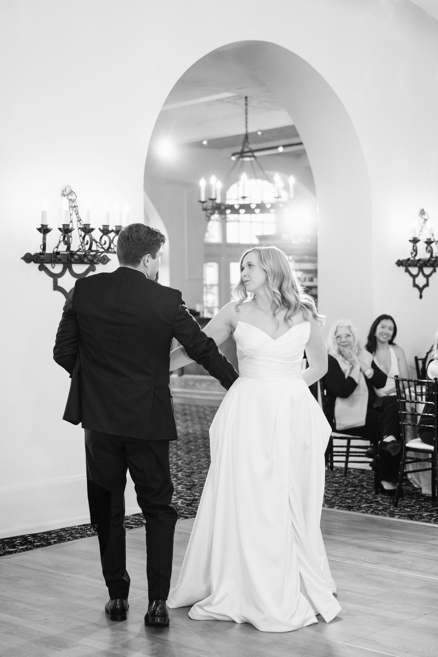 A couple shares their first dance at an elegant reception venue with arched doorways and ornate chandeliers.