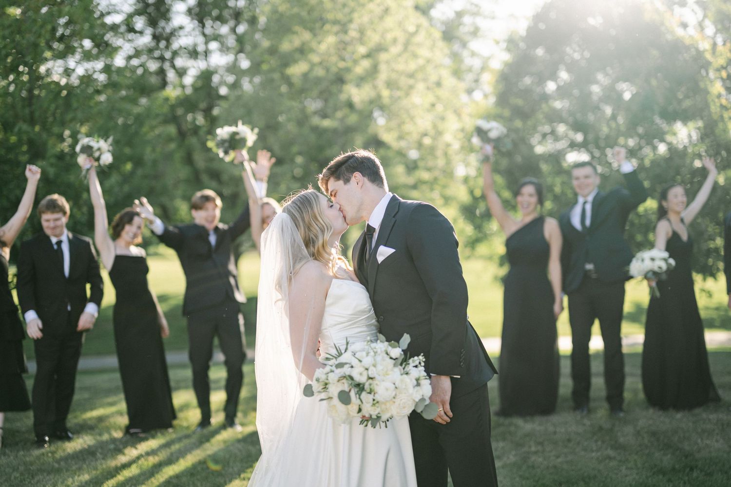 A wedding party gathered on an outdoor patio surrounded by lush green trees on a bright sunny day.