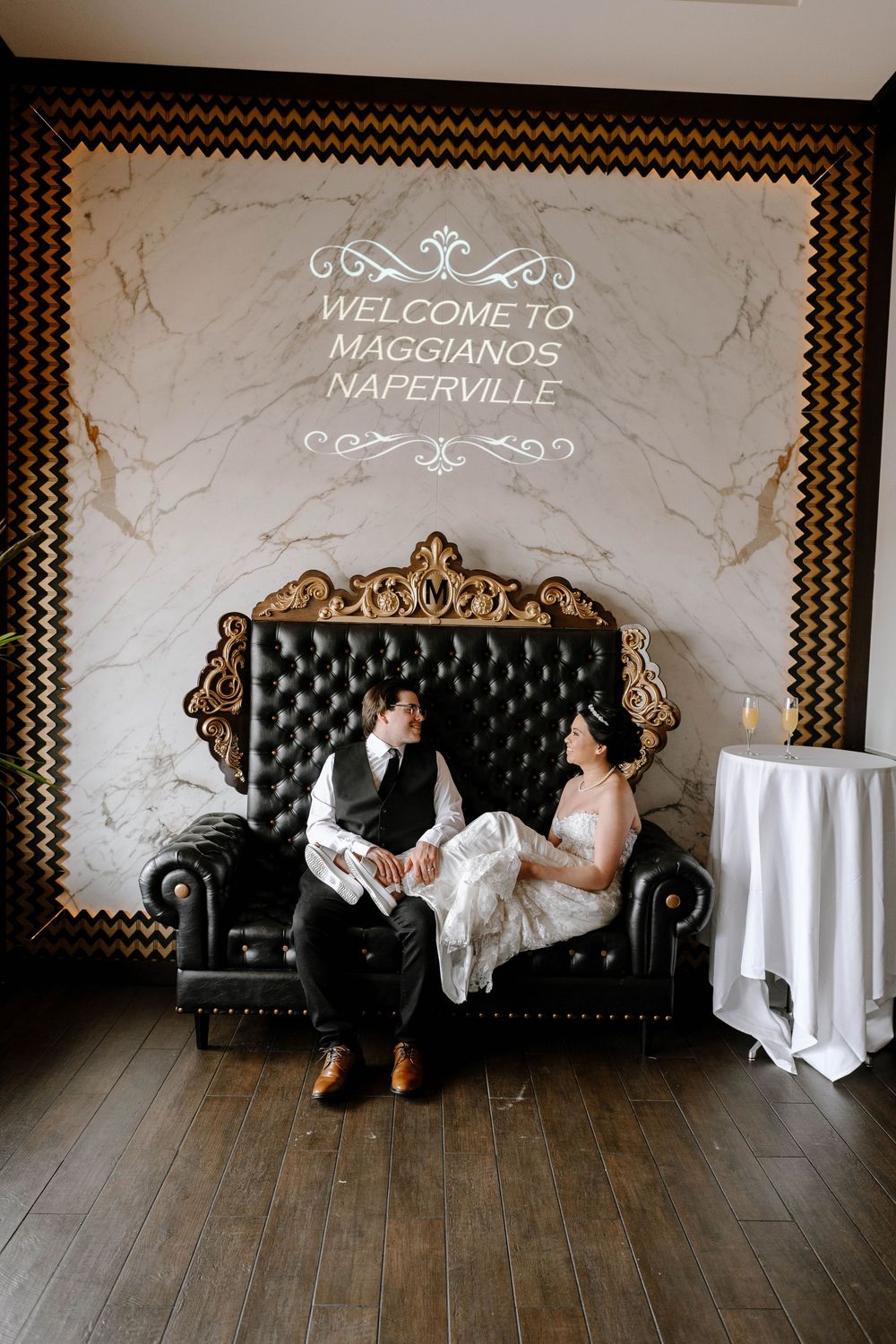 A wedding couple poses casually on an elegant black tufted sofa beneath decorative signage.