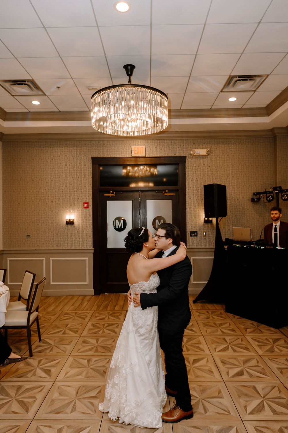 A couple shares their first dance under an elegant crystal chandelier in a ballroom with patterned flooring.
