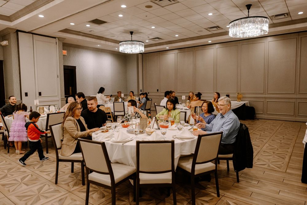 Reception guests seated at round tables in an elegant ballroom with crystal chandeliers and beige walls.