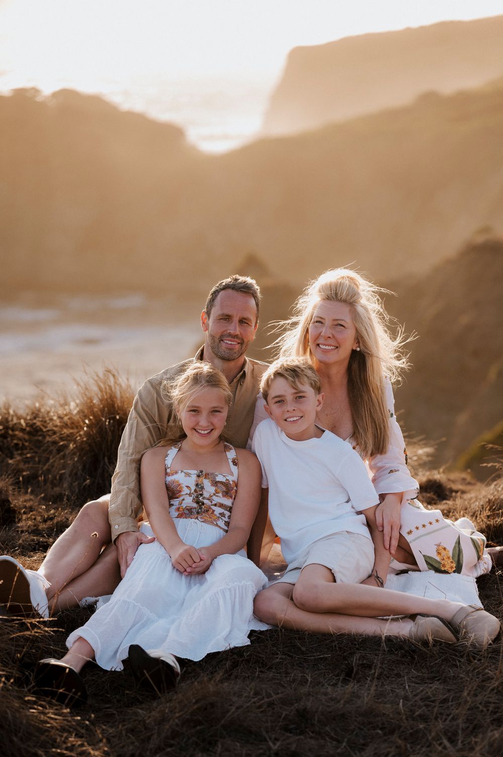 A family sits together on a hillside during golden hour with mountains and scenic landscape in the background.