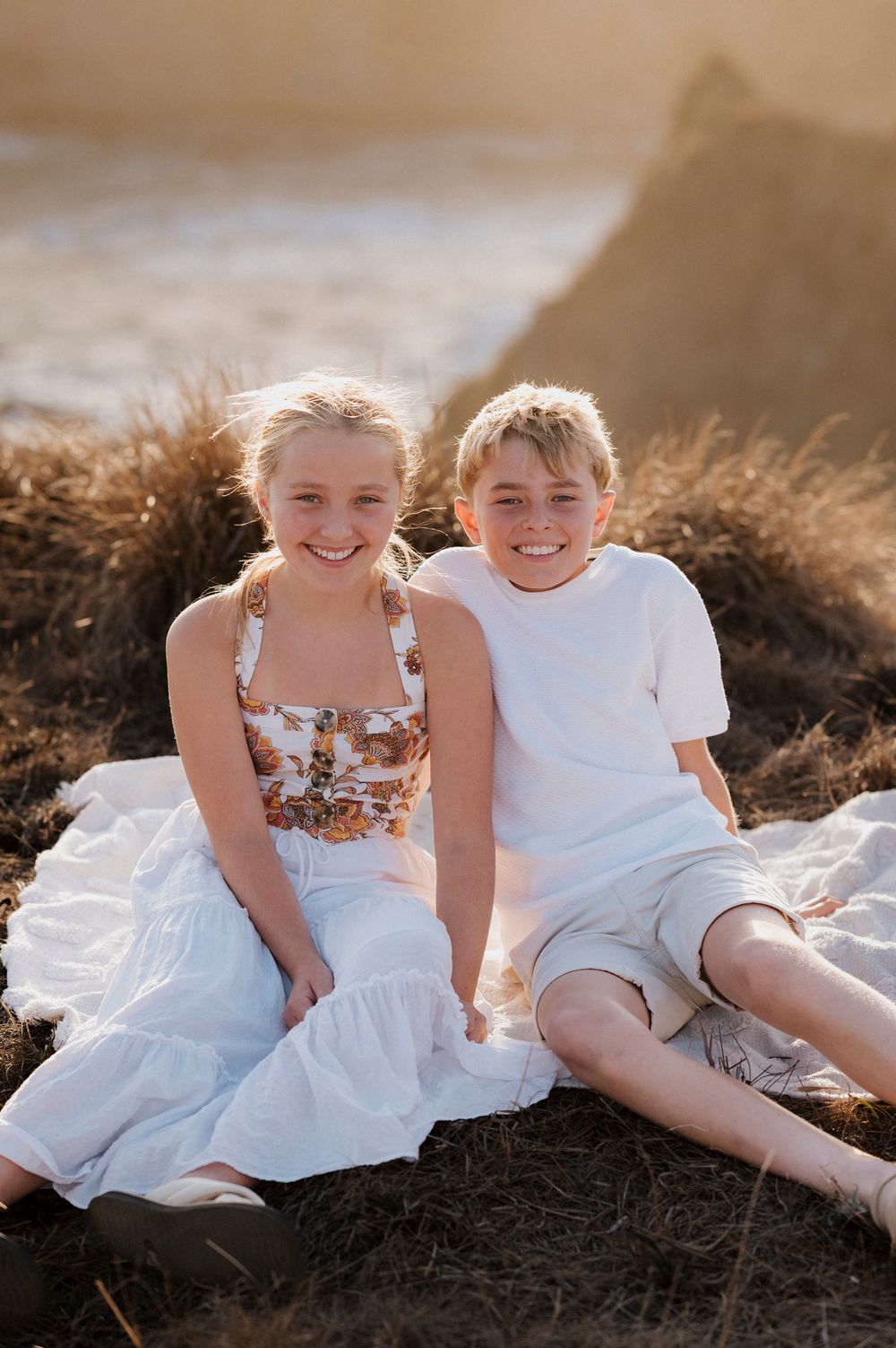 Two children in white summer outfits sit together smiling on a sunny hillside with dry grass in the background.