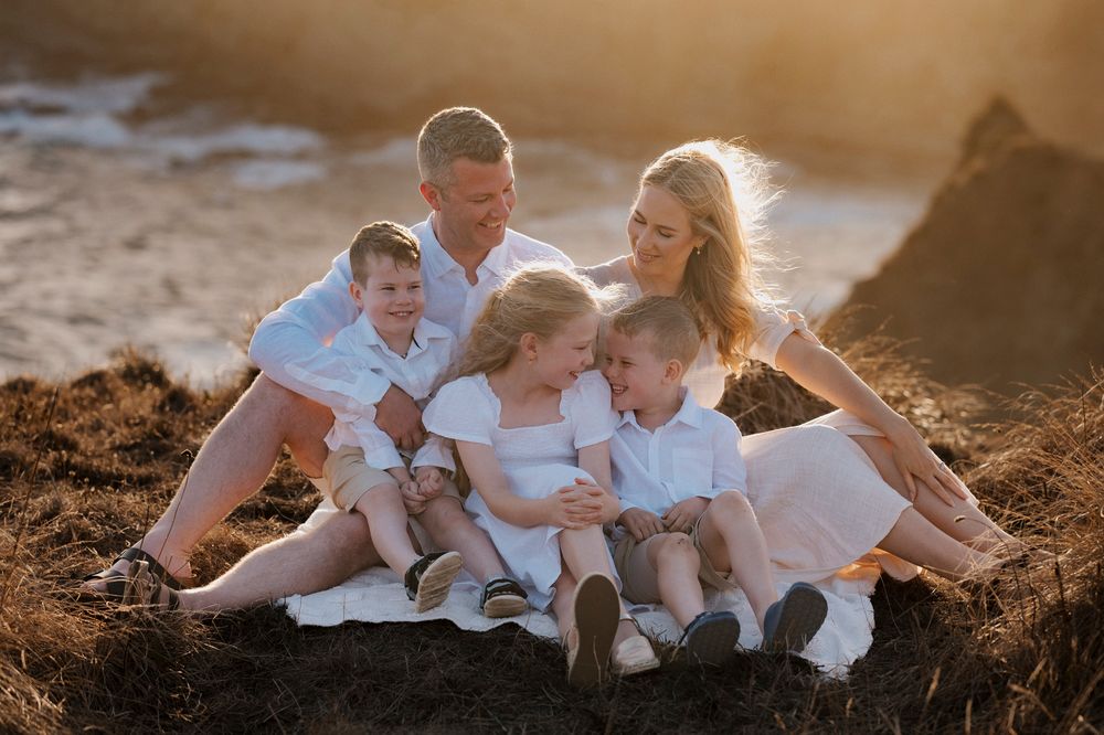 A family wearing white clothes sits together on a cliff at sunset creating a warm and intimate portrait.