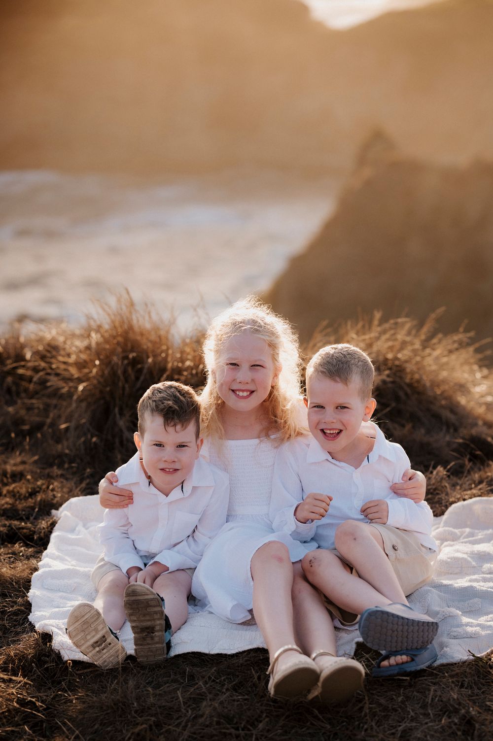 Three siblings in white outfits sit together on a blanket at sunset, smiling and enjoying a warm outdoor moment.