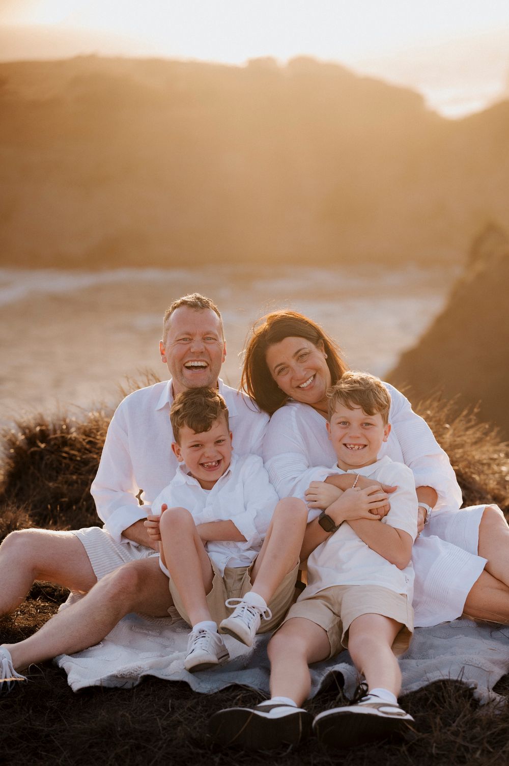 Family shares joyful laughter while sitting together on blanket during golden hour sunset at scenic overlook.