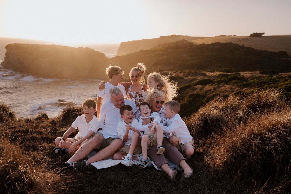 A group in white outfits sits together on coastal cliffs at golden hour as sunlight bathes the scenic ocean landscape.