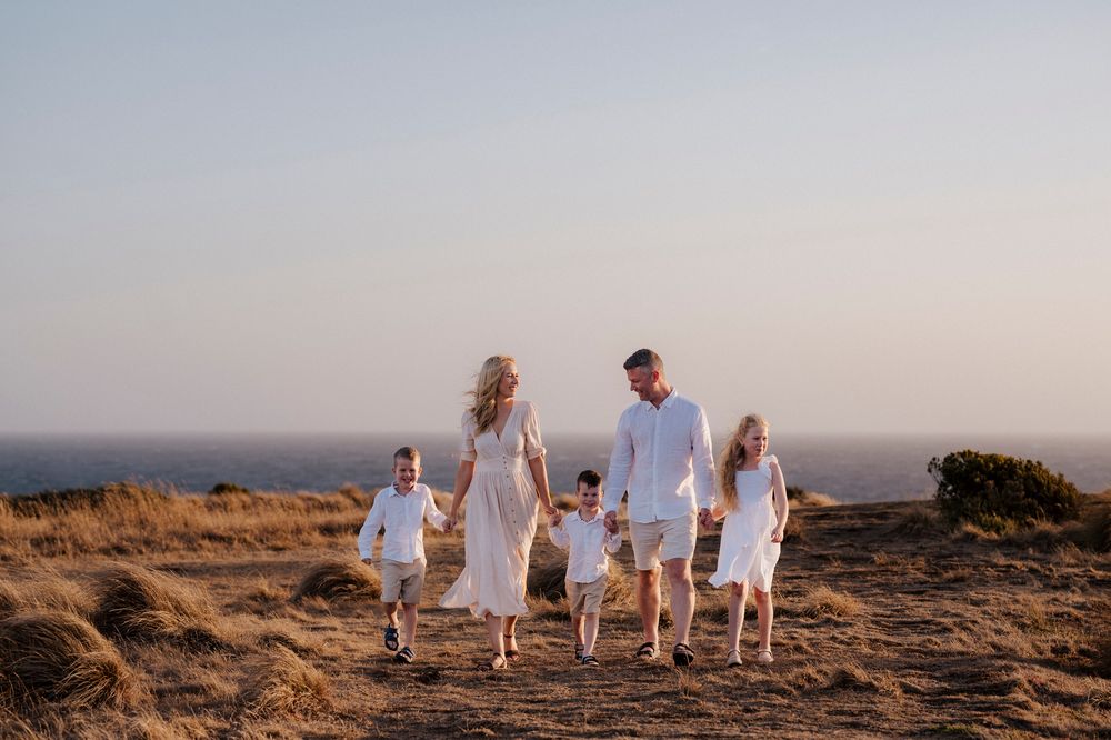 A family holding hands walks together on a scenic hilltop during sunset in white summer outfits.