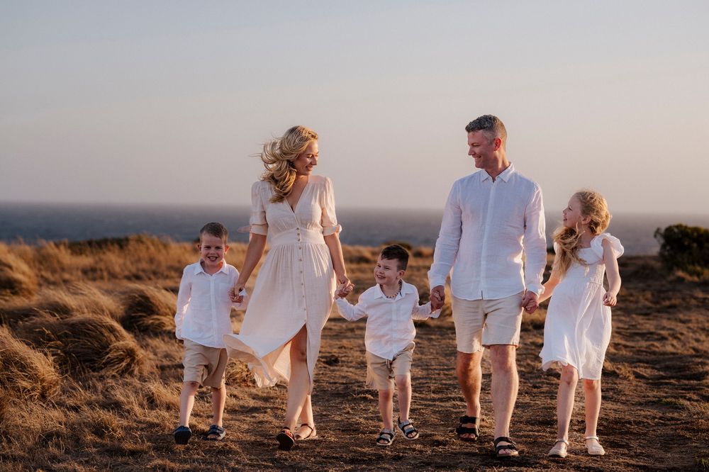Family of five wearing coordinating white outfits walks together on a hilltop at sunset against ocean backdrop.