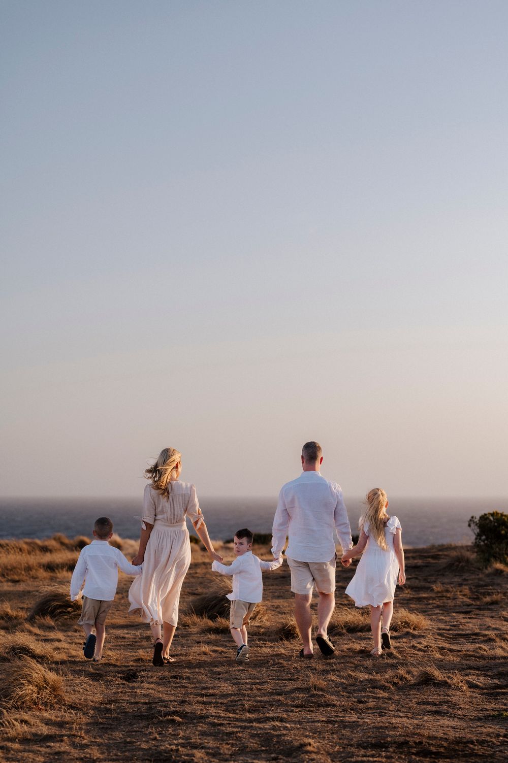 A family in white outfits walks together along a hilltop path at sunset against a misty sky background.