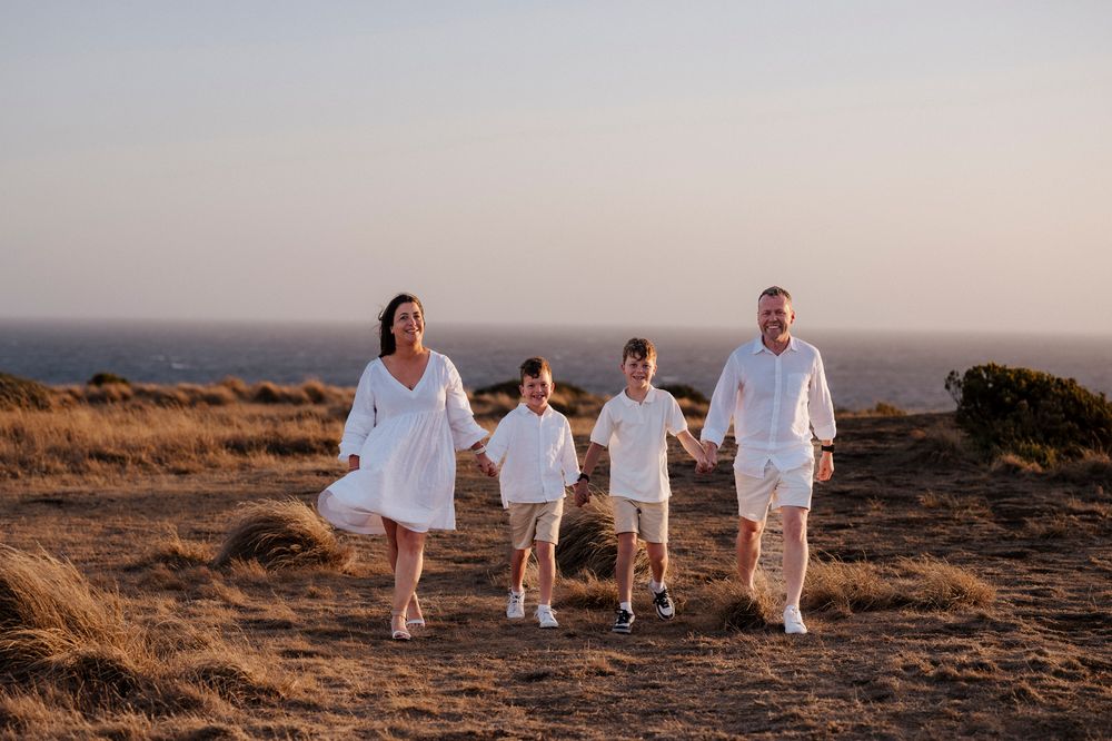 A family wearing white outfits walks together on a hilltop path at sunset with an ocean view in the background.