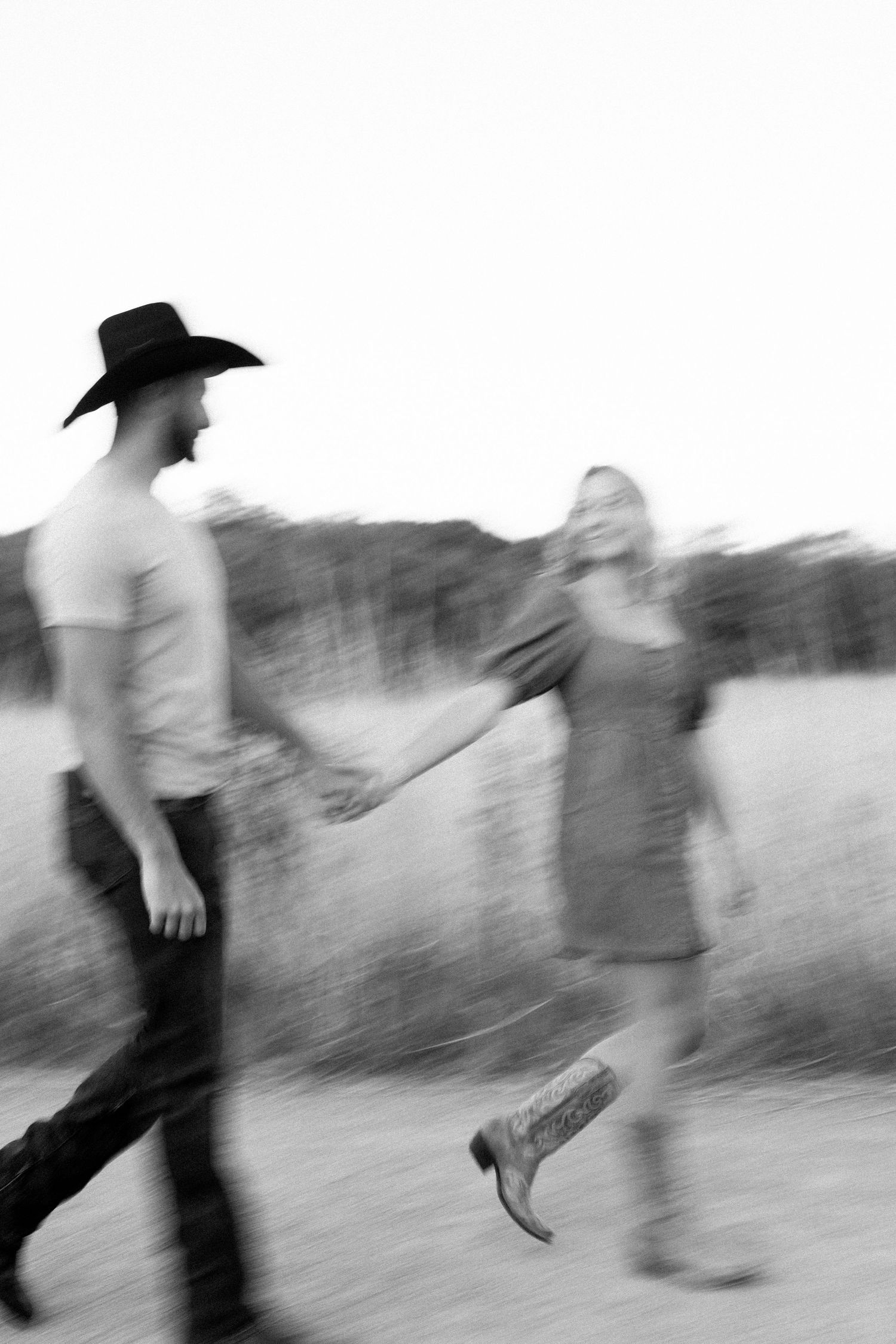 A blurred black and white motion photo of two people walking together outdoors with one wearing a cowboy hat.