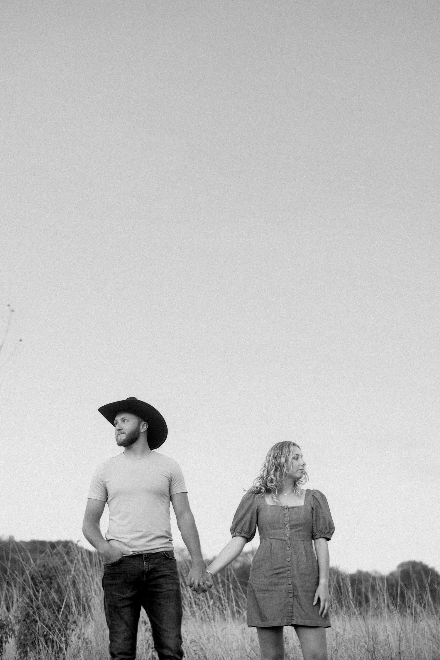 Black and white photo of a couple holding hands in a field, with the man wearing a cowboy hat and the woman in overalls.