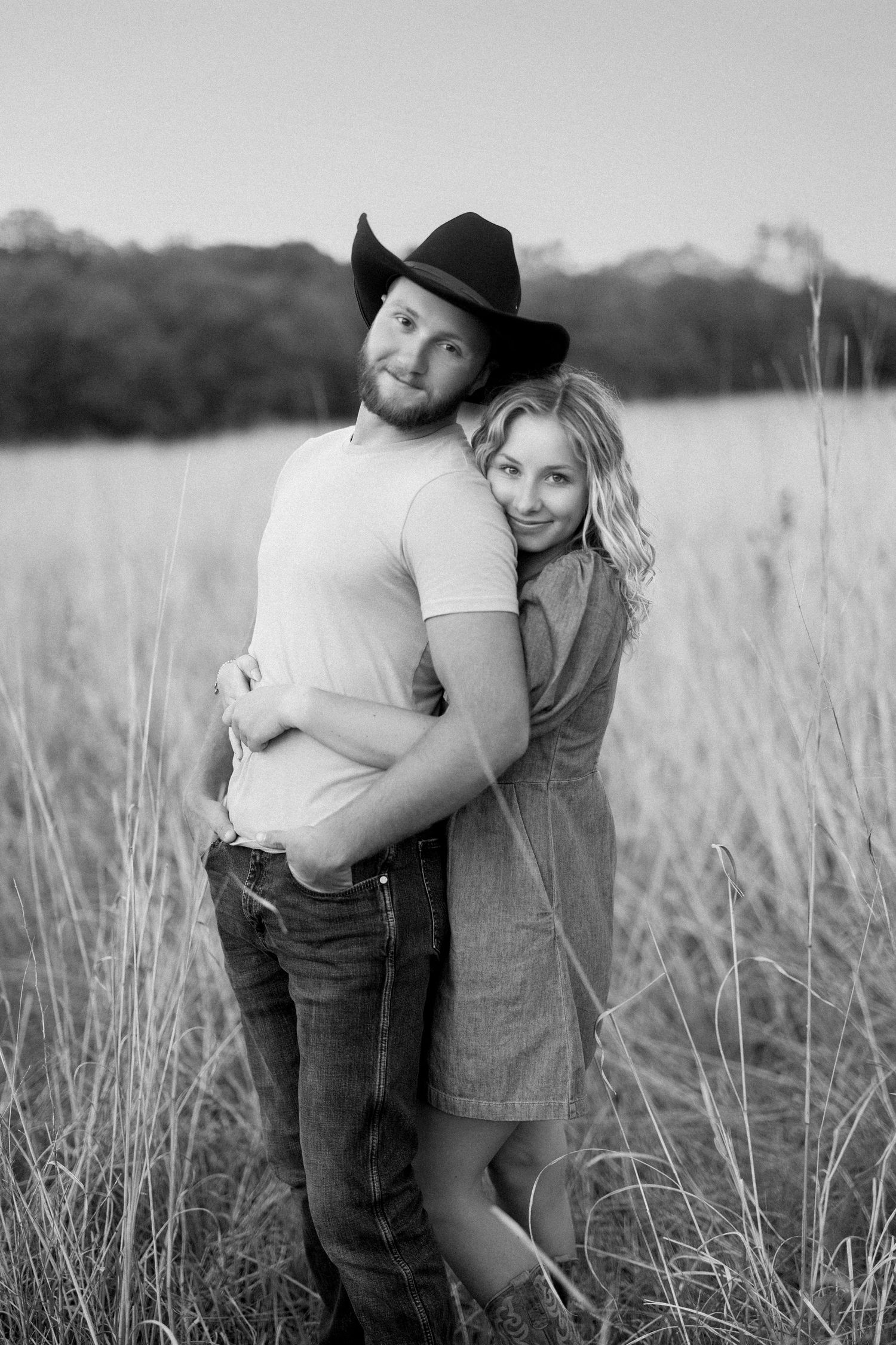 A couple embraces in a sunlit field of tall grass, captured in black and white.