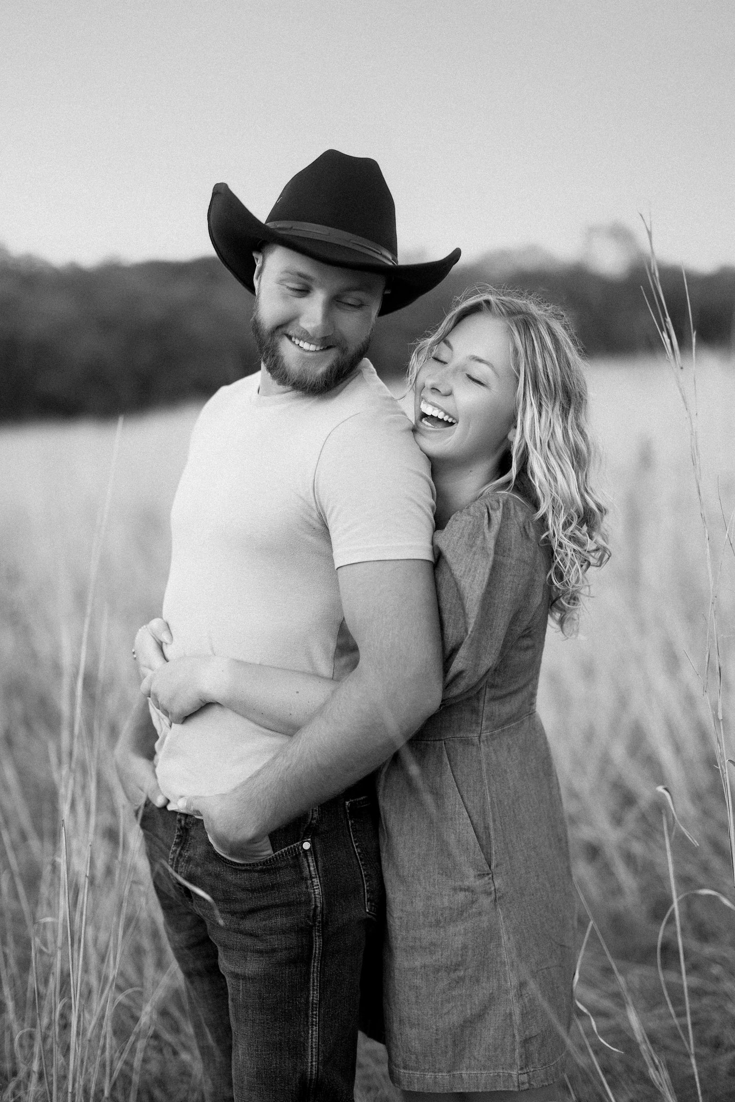 Black and white portrait of a joyful couple embracing in a tall grass field outdoors.