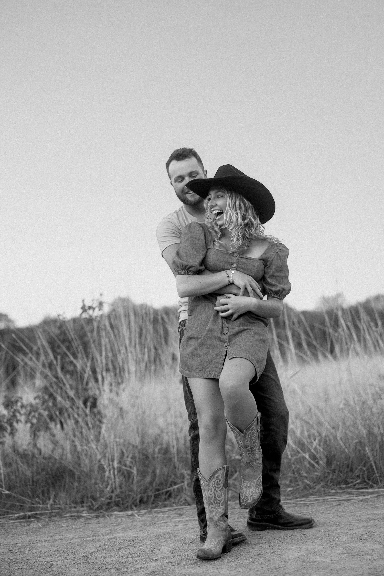 A couple shares a playful embrace on a dirt path surrounded by tall grass in black and white photography.