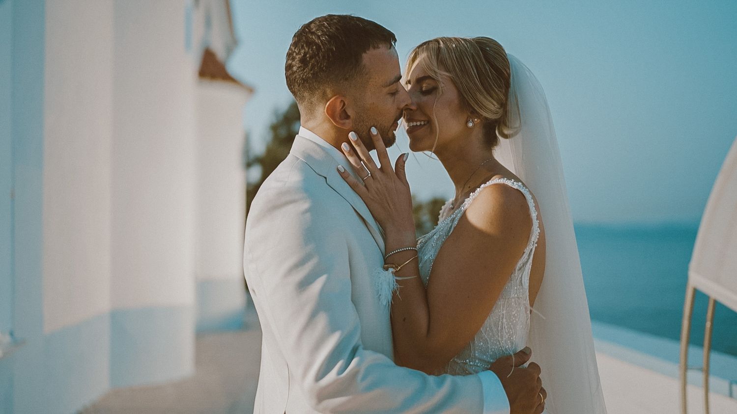 A romantic wedding couple embraces intimately against a bright blue Mediterranean backdrop on a sunny day.