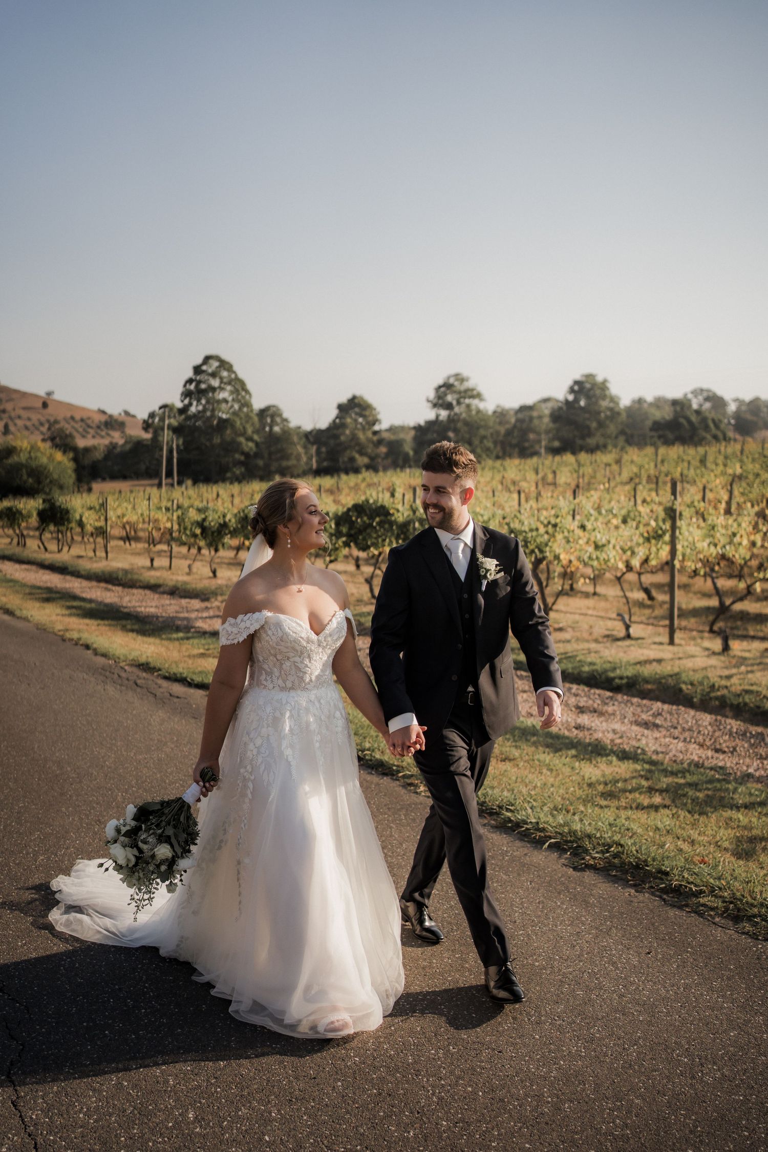 A couple walks hand in hand along a path through a scenic vineyard.
