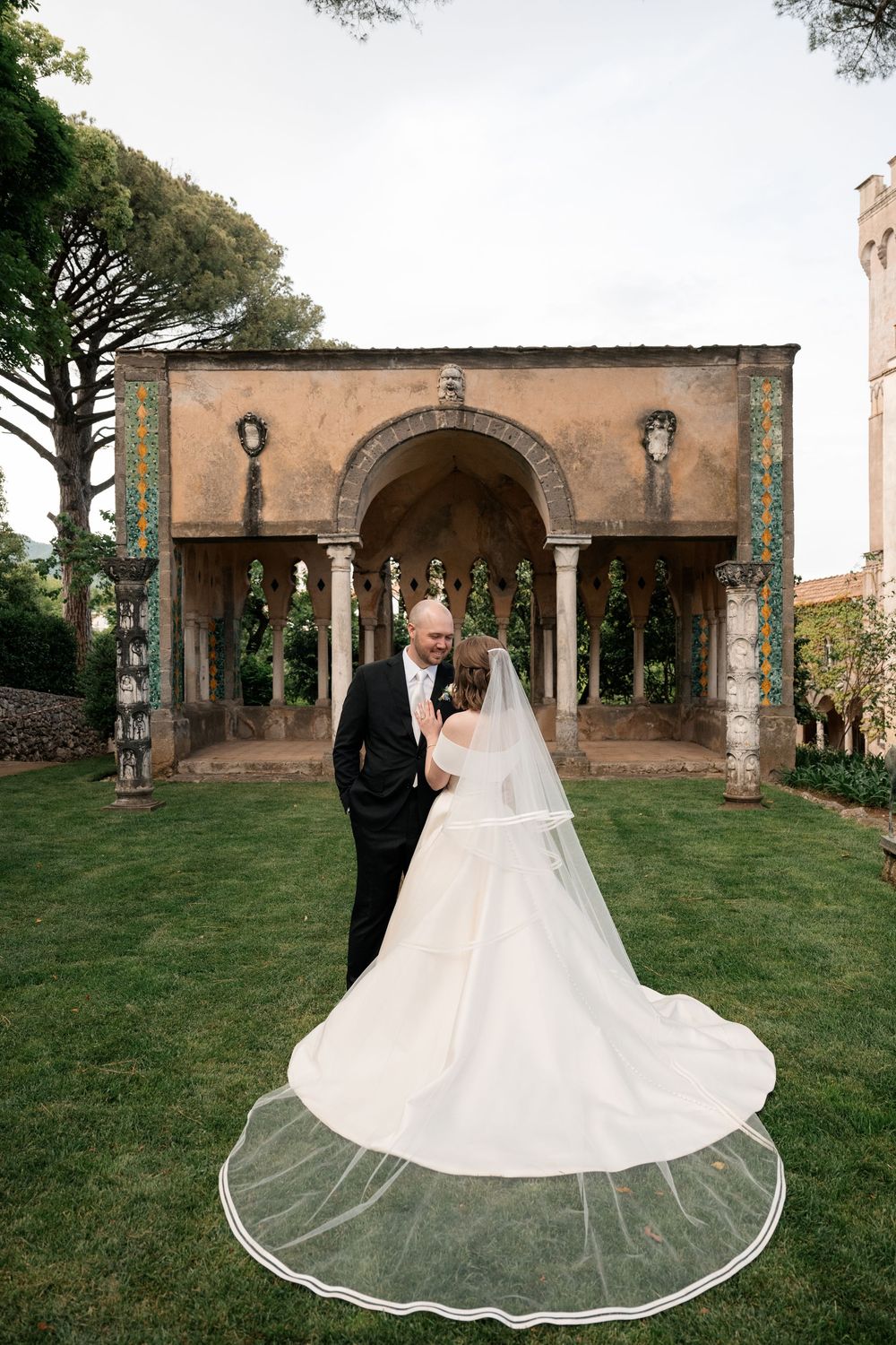 An elegant wedding couple poses beneath stone arches in a romantic garden setting.