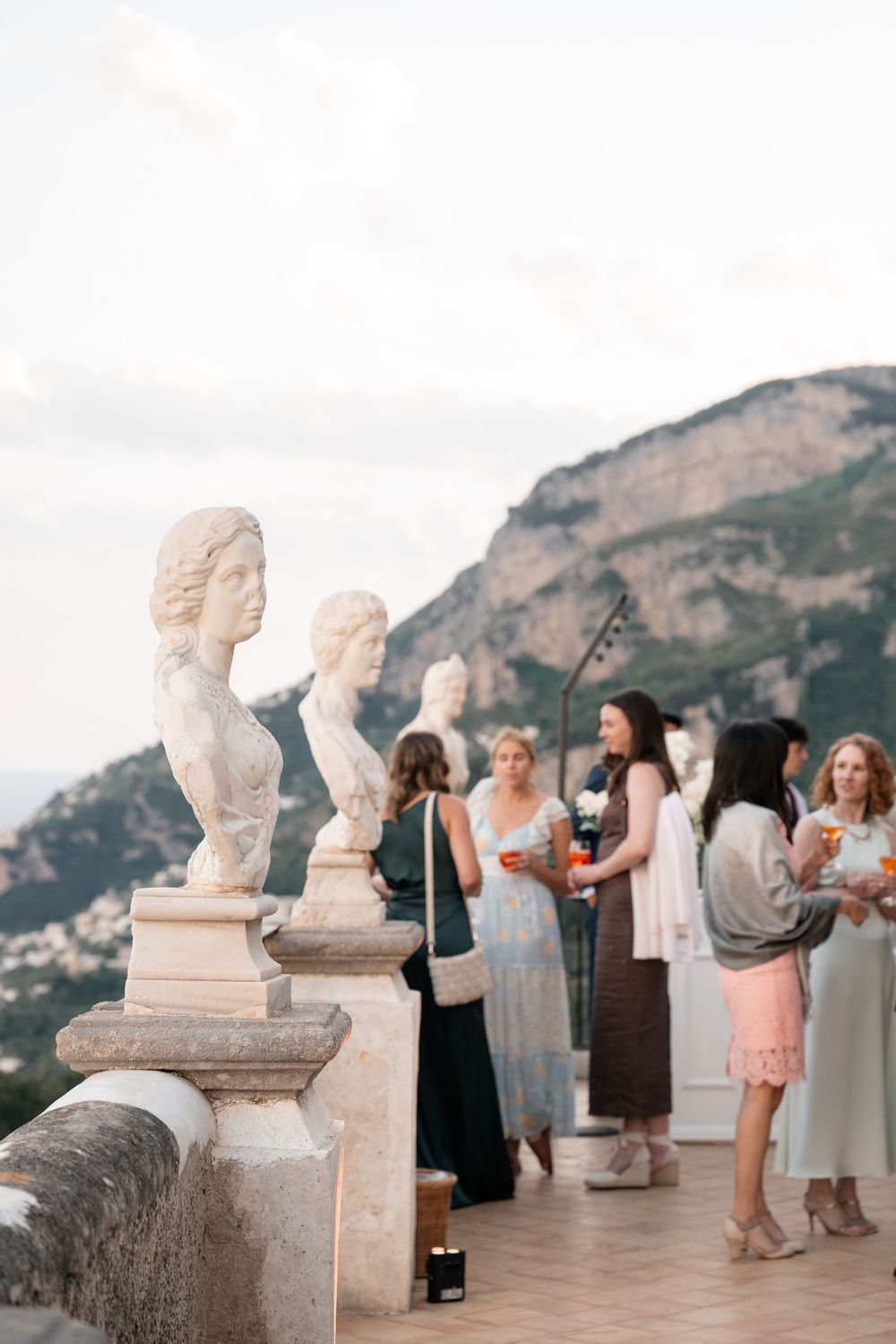 Marble busts on a balcony railing with scenic mountain views and guests gathering at an elegant outdoor event in Italy.