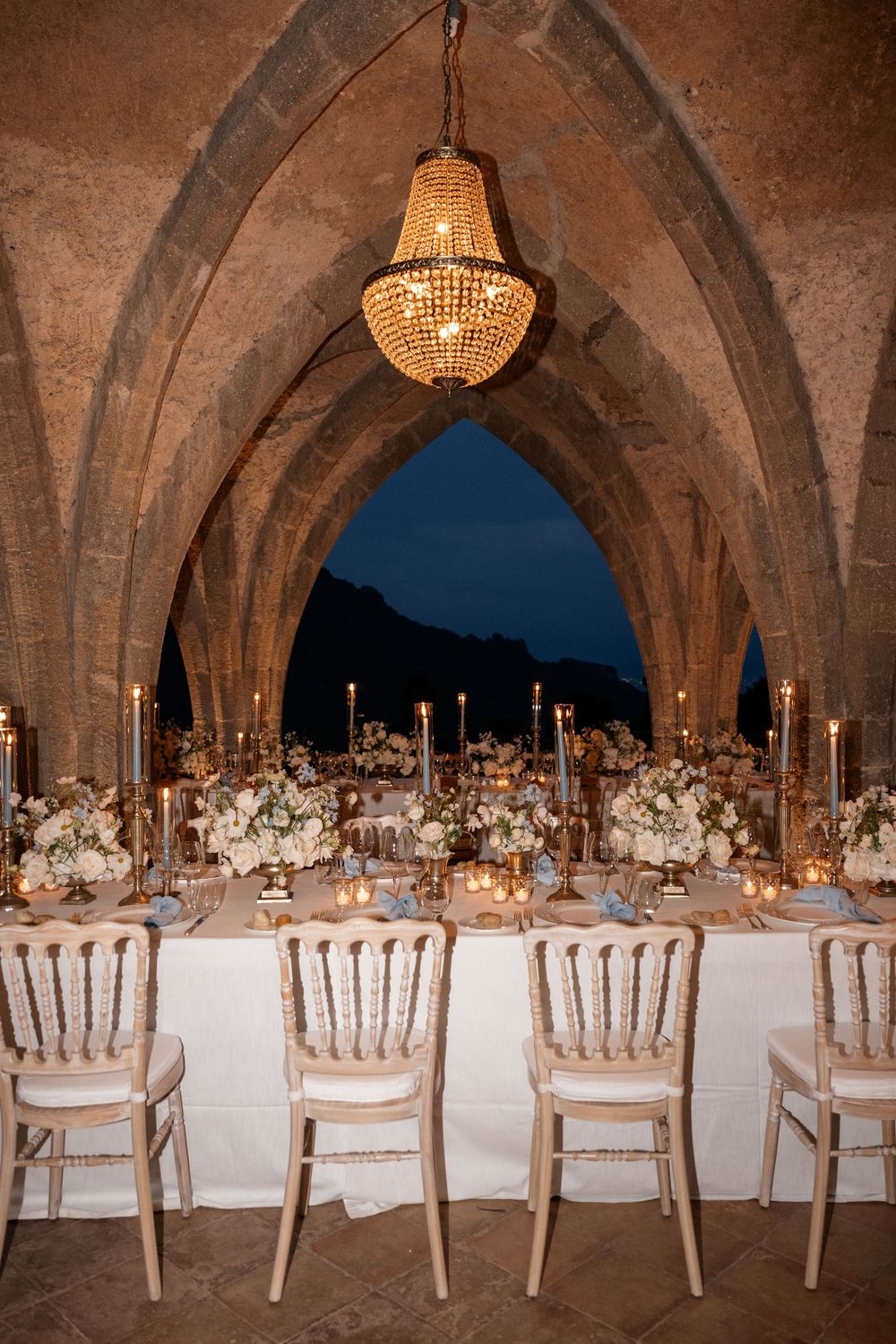 Gothic stone archway decorated with crystal chandelier and elegant table settings at dusk for an outdoor wedding reception.