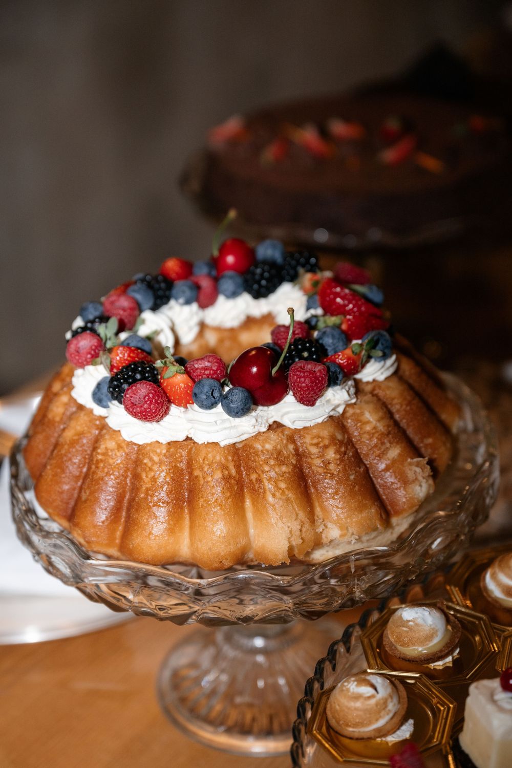 Bundt cake topped with fresh mixed berries and whipped cream displayed on an ornate glass pedestal.
