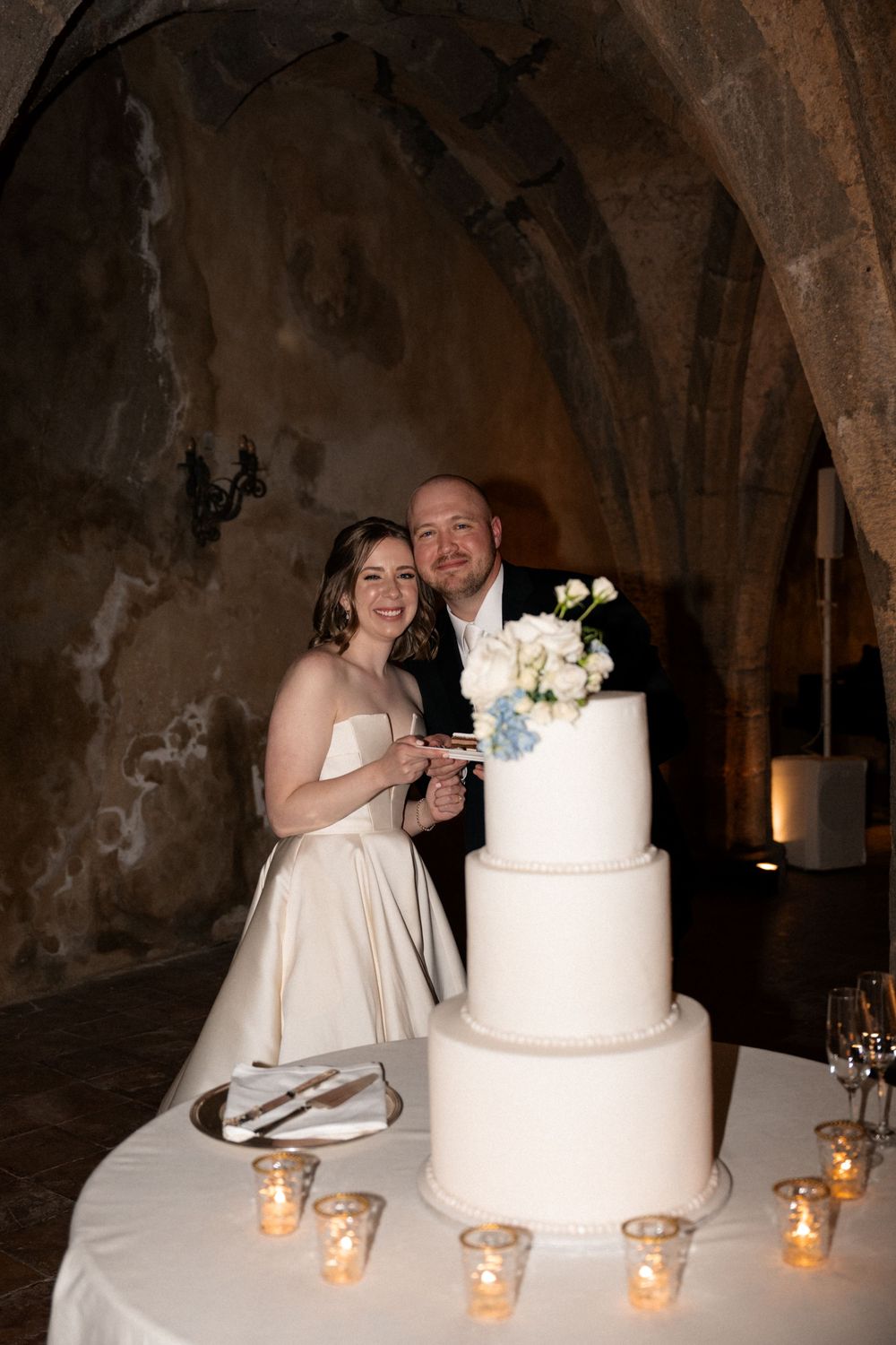 Couple cutting their elegant white wedding cake decorated with flowers while candlelit tea lights line the table.