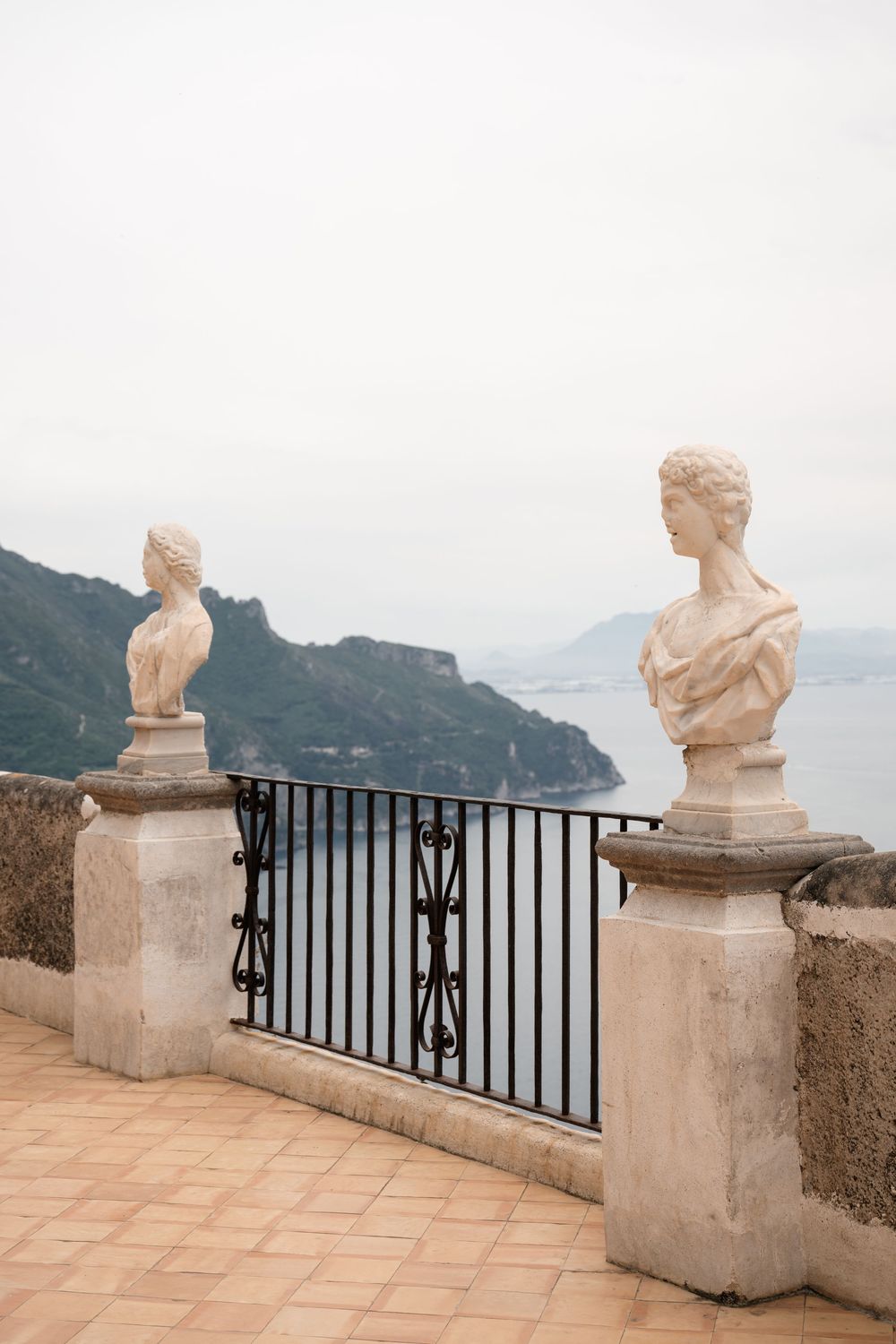 Two classical stone busts on balcony overlooking coastal mountain views.