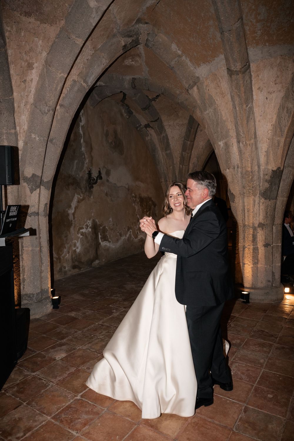 Couple dancing in formal attire under historic stone arches lit by ambient lighting.