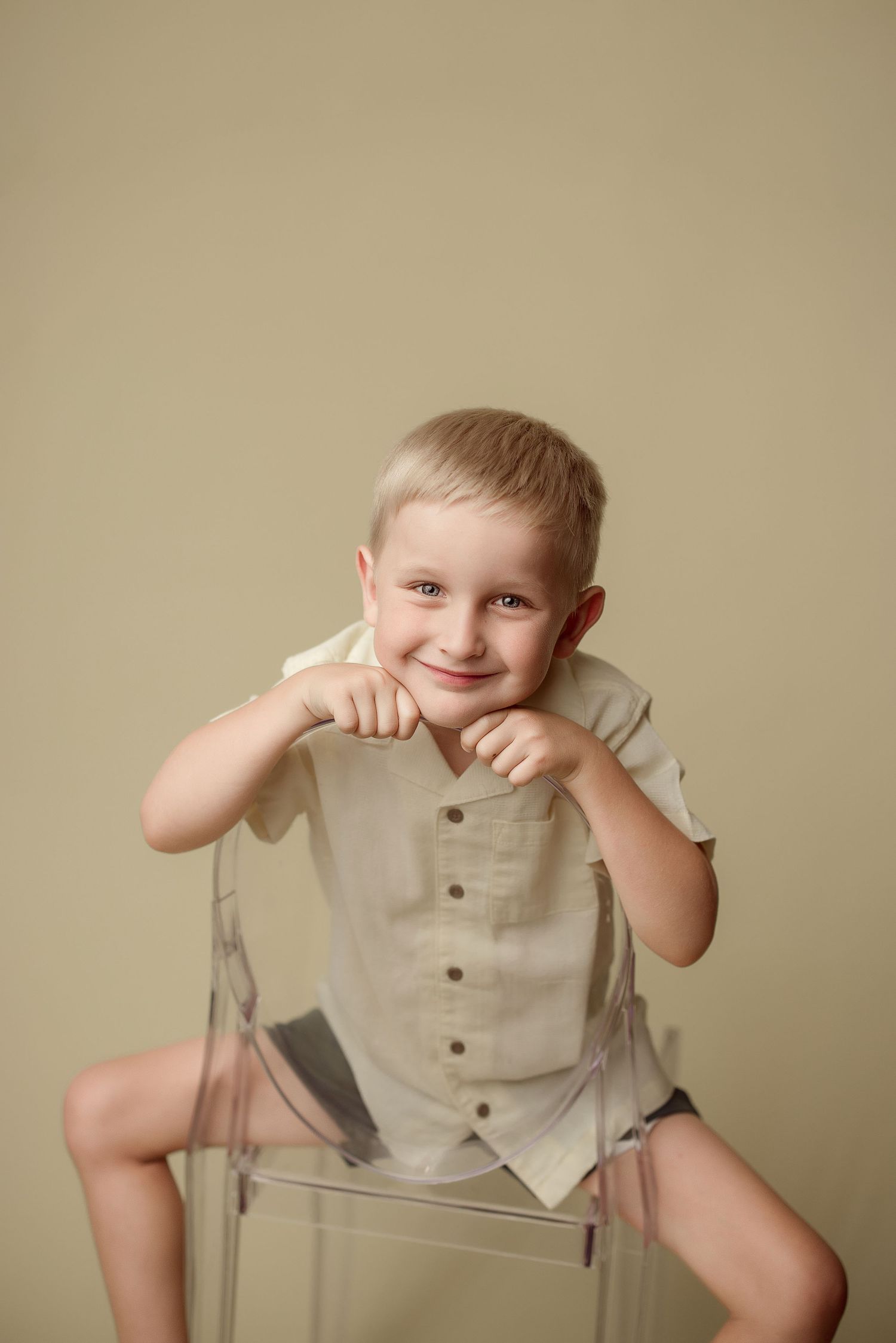 Small child wearing beige button-up shirt poses playfully while sitting on clear acrylic chair against neutral background.