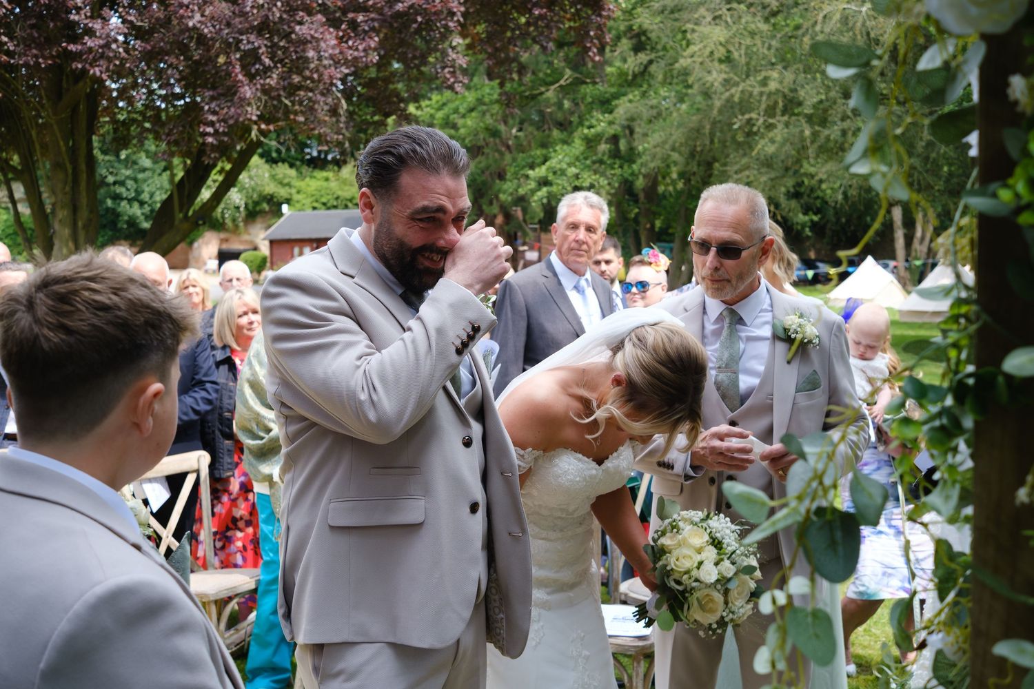 Bride had arrived at the front of the aisle with her dad. Both bride and groom are emotional and crying. Bride is leaning forwards.