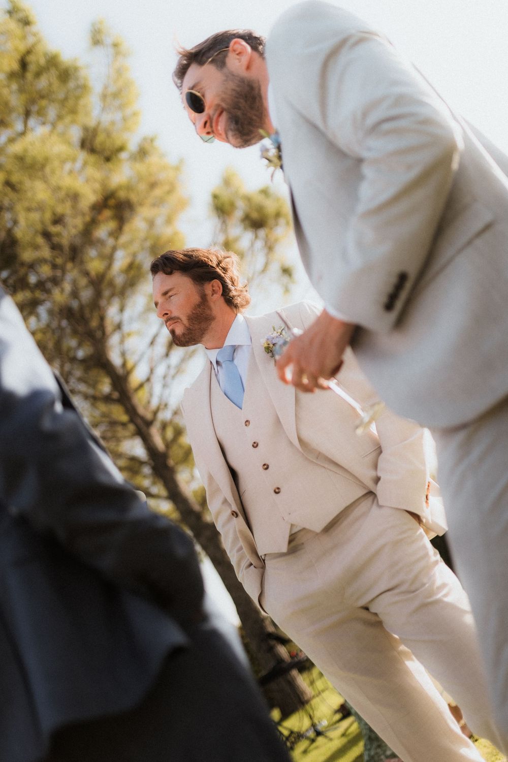 People in light colored formal attire gather for outdoor wedding photos in natural sunlight.